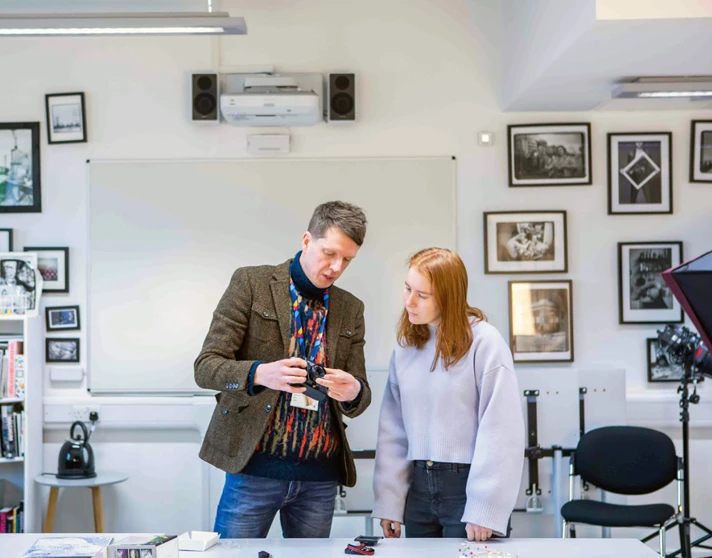 Lecturer demonstrating camera functions to a student in a photography classroom filled with artwork. Lecturer demonstrating camera functions to a student in a photography classroom filled with artwork.